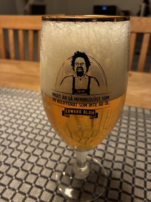 Ett glas öl med skum, tryck av en man och text om kolhydrater. Glaset står på en mönstrad duk.
