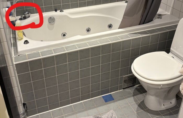 Badkar med blandare markerad i rött, ansluten slang syns under kanten. Toalett intill.