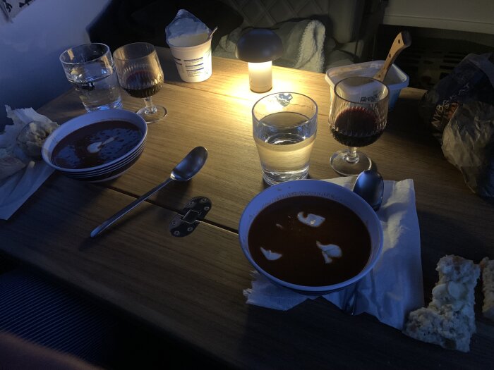 Skål med gulasch på bord, omgiven av vinglas, vatten, sked, bröd och margarin, belyst av en lampa i skärgårdsmiljö.