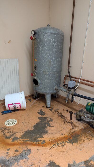 Urtappning av rostig metalltank på golv med vattenpöl och verktyg bredvid vitt plasthink.