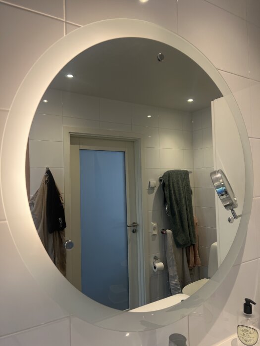 Rund badrumsspegel med integrerad belysning ovanför ett handfat, omgiven av vit kakel. Handdukar hänger på väggen i spegeln.