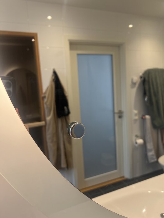 Rund spegelknopp i badrum med synlig dörr och hängande handdukar i bakgrunden.