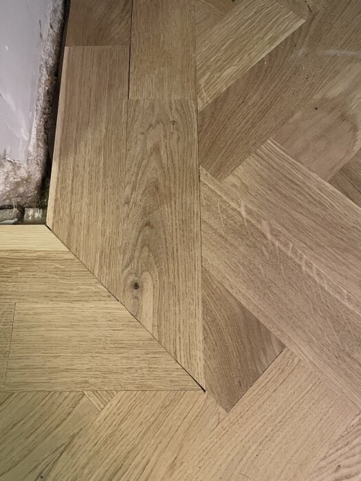 Närbild på fiskbensparkettgolv med synliga springor mellan stavarna och en saknad triangelbit vid väggen.