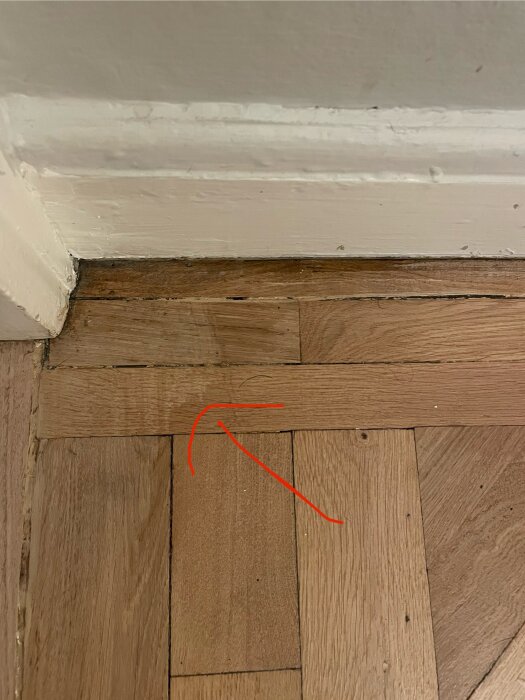 Fiskbensparkett med glipor och en saknad triangel mot listbandet vid väggen. Röd pil markerar ett problemområde.