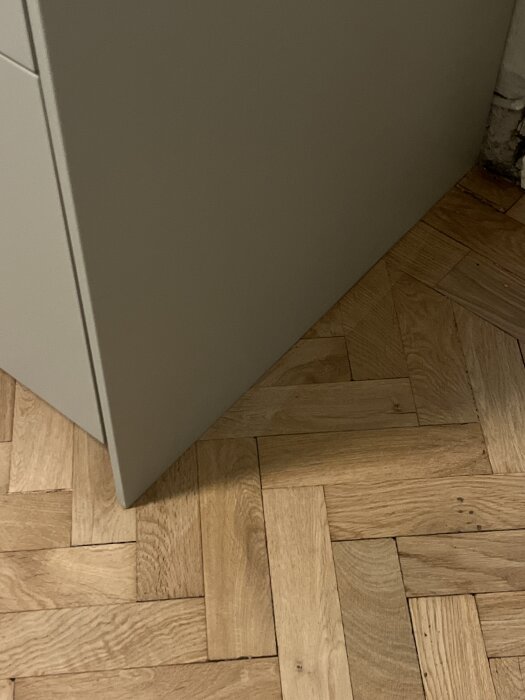 Fiskbensparkett med glipor och en ojämn övergång till ett annat rum; nära en vit vägg. Skador och slipspår syns på golvet.