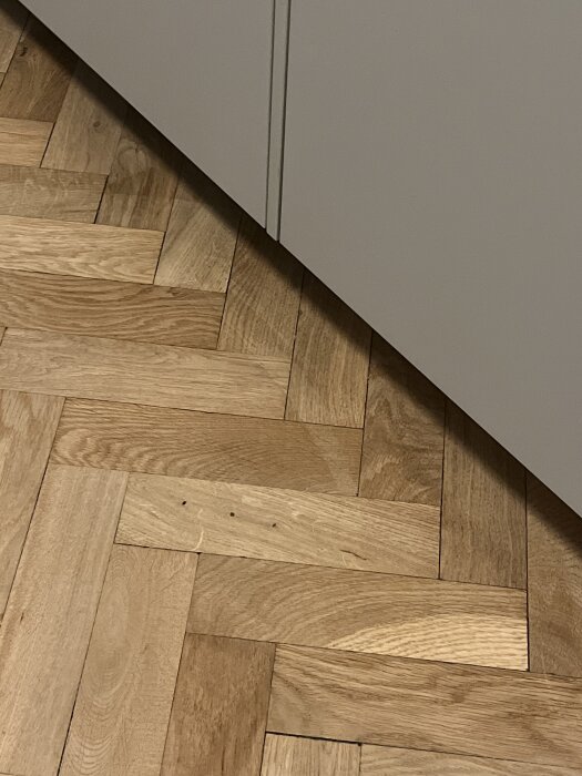 Fiskbensparkett med synliga glipor och trasiga bitar nära en skåplucka.