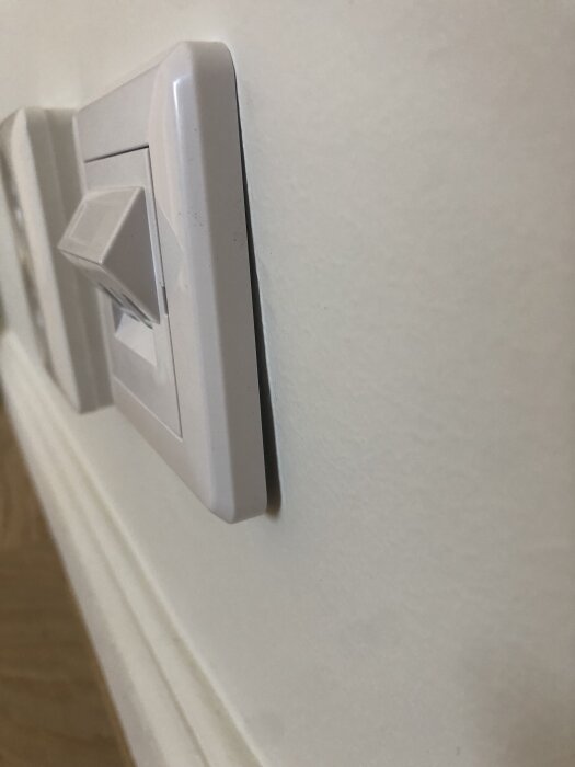 En vit strömbrytare på en vägg där ramen verkar lossna lite från ytan.
