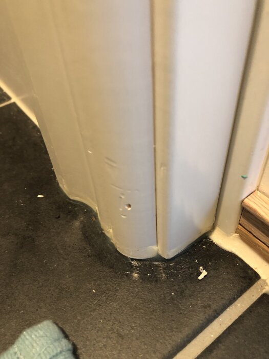 Skadad golvlist i ett hörn med märken och hål, placerad på mörkt klinkergolv nära en dörrkarm.