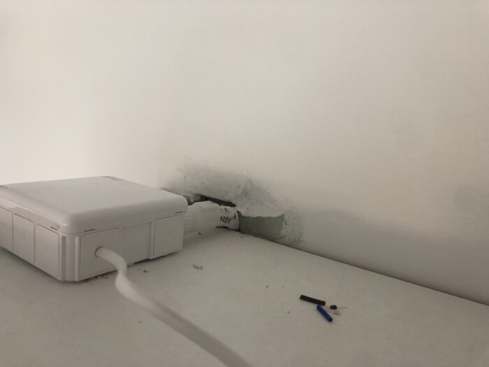 Vit vägg med sladd och hål, vit dosa på hylla, mindre delar på ytan.