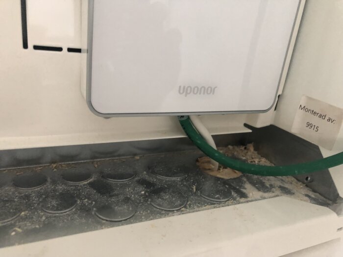 Närbild av en monterad Uponor-enhet med en grön kabel och smuts eller damm på panelen nedanför, samt en etikett med texten "Monterad av: 9915".