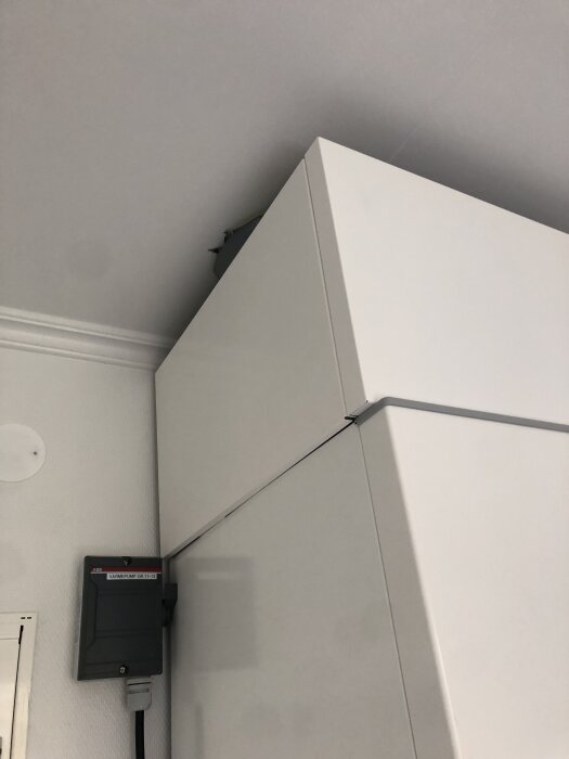 Övre delen av ett högt, vitt skåp under ett tak med vit taklist, bredvid ett elskåp märkt "Värmepump GR11H-2".