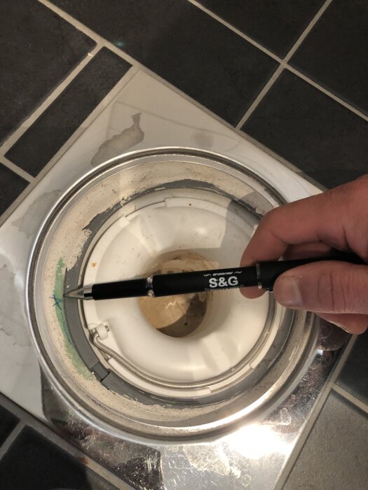 Hand pekar med penna på duschbrunn med missfärgning på klinkergolv.