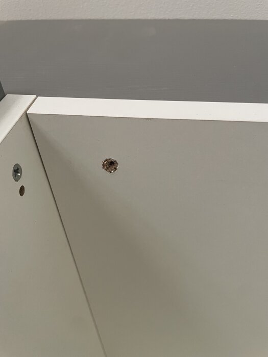 Närbild på ett skåphörn med en stor och en liten borrhål i en vit yta, monterad med skruvar i en mörkare yta.