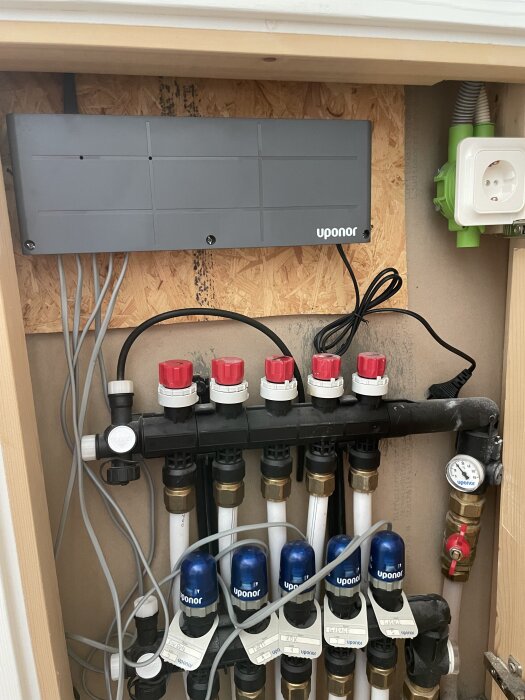 Grå Uponor-central med flera anslutna rör och ventiler, nätadapter och strömuttag synliga bakom centralen.