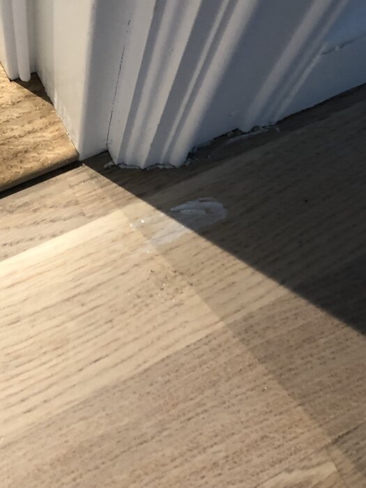 Dörrkarm mot laminatgolv visar användning av fogmassa eller spackel för att fylla en glipa, med ljus och skugga på golvytan.