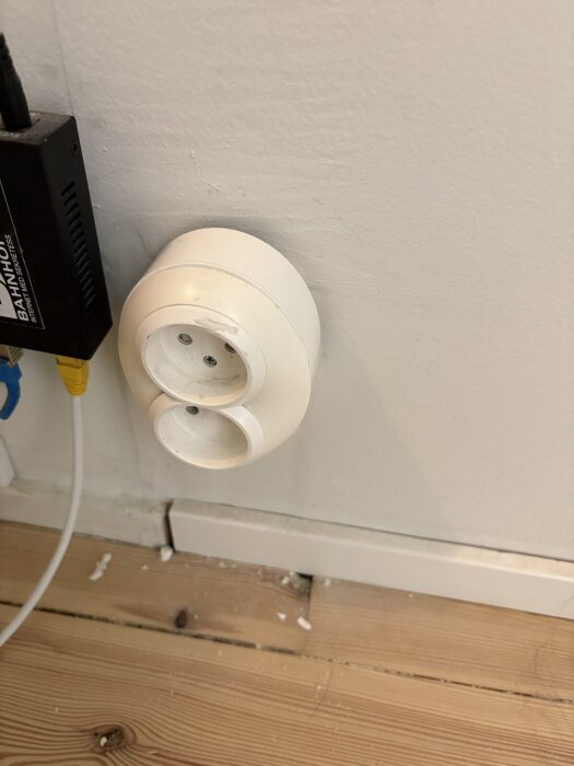 Dubbeluttag med förhöjningsring på väggen, övre uttag fungerar inte korrekt. Golvbräda och sladdar syns i bilden.