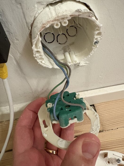 En hand håller en grön insats från ett eluttag med synliga kablar i en ojordad dosa i väggen.
