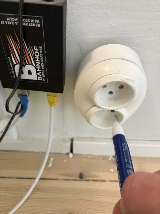 Ojordad vägguttag med en spänningsprovare i det nedre uttaget, internetbox till vänster, träpanelgolv.