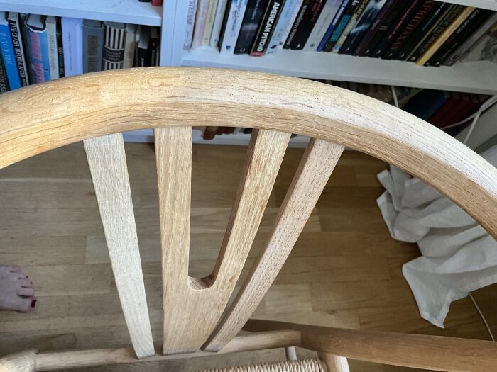 Ryggstöd på en trästol, eventuellt av ask, mot en bakgrund med bokhylla och trägolv.