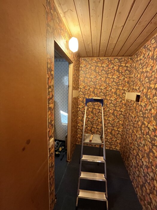 Trångt rum med blommig tapet, en stege och öppning i väggen där en dörr tidigare suttit. 70-talsstil med träpanel i taket.