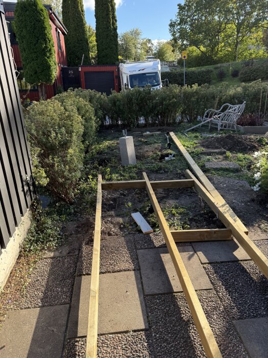 Trädgård med pågående altanbygge, reglar över plattor, buskar i bakgrunden, och en husbil parkerad bakom en häck.