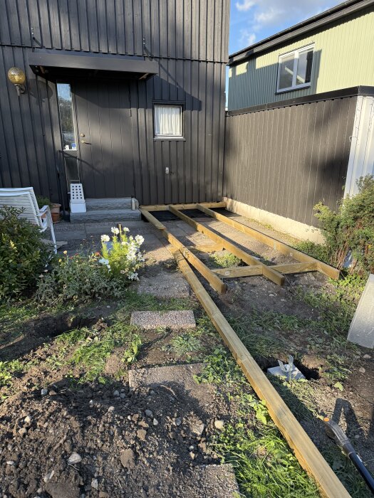 Regelram för altan byggs på en lutande upphöjd tomt nära ett svart träklätt hus, med blommor och buskar i bakgrunden.