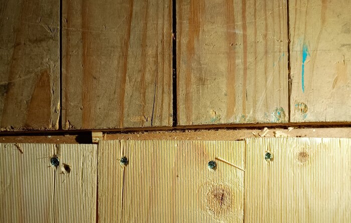 Trägolv där brädorna är ojämna och provisoriskt skruvade efter en reparation, med synliga springor och skavanker i träytan.