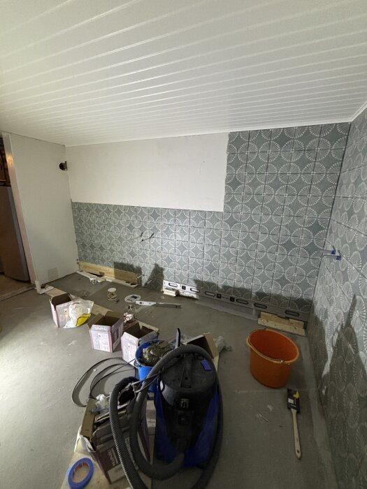 Vitt rum under renovering med halvfärdig kakelvägg. Verktyg, kartonger och en dammsugare på golvet.