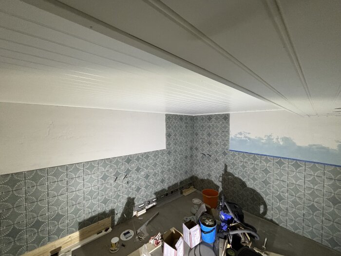 Rum under renovering med väggar delvis kaklade i grått mönster, vita paneltak och verktyg synliga på golvet.