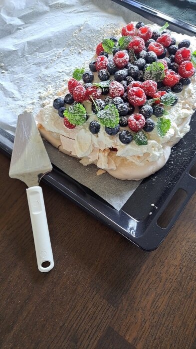 Pavlova toppad med bär och pudrad med florsocker på en bakplåt, bredvid en vit tårtspade.