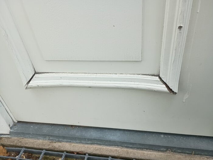 Dekorlister på vit ytterdörr, delvis lösa och slitna, med färg som skavts av, synliga längs nedre och högra kanterna av dörrpanelen.