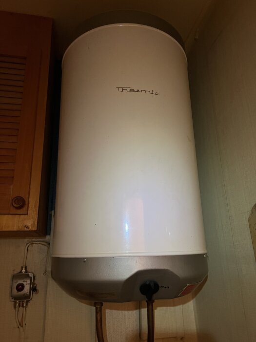 Gammal varmvattenberedare från 70-talet med en kran och kopparrör synliga, placerad i ett träskåp.