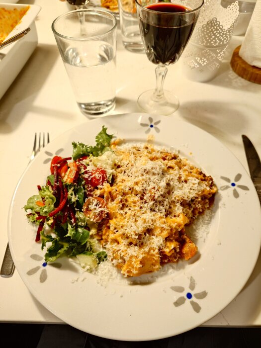 Lasagne med ett lager parmesan och sallad på en vit tallrik, med ett glas rödvin och ett glas vatten bredvid.