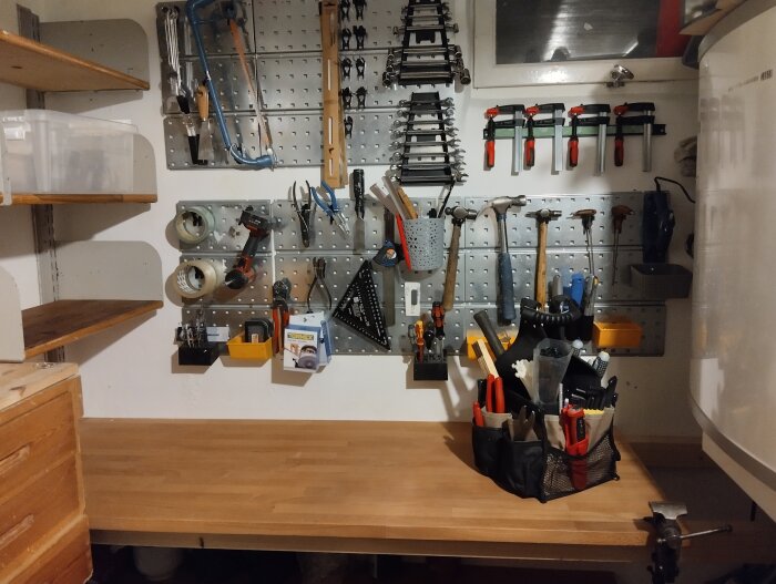 Verktygstavla med diverse verktyg som tänger, skruvmejslar och nycklar, samt en arbetsbänk med verktygsväska i ett organiserat förråd.