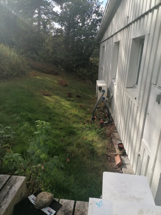 Gräsmatta med sluttning mot en vit träfasad på ett hus, stenar vid husgrunden, filter och rör synliga, omgiven av träd och buskar.