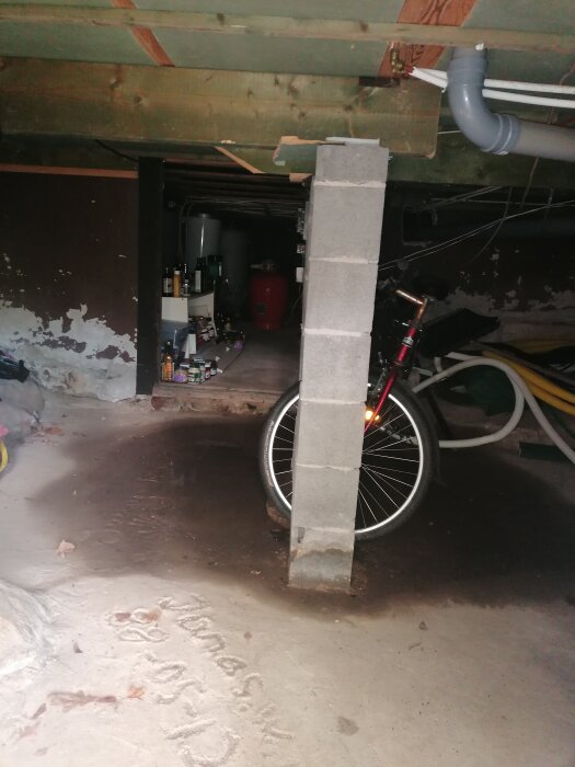 Källare med synliga fuktfläckar på golvet, en cykel, hyllor med flaskor och rör i taket. En betongpelare mitt i bilden.