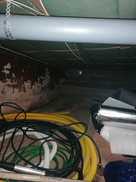 Utrymme under ett hus med olika färgglada rör och slangar, en stor grå ventilationsrör i taket och delvis skadade väggar.