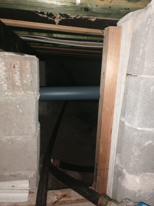 Betongväggar med rör i källarutrymme under huset. Öppning med träbeklädnad och en del mörkt fuktmärke synligt ovanför.