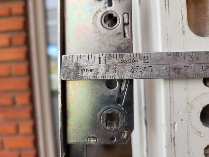 Mätning av dornavstånd med linjal på metallbeslag för spanjolett bredvid dörrkarm, fokuserad på byggprojekt.