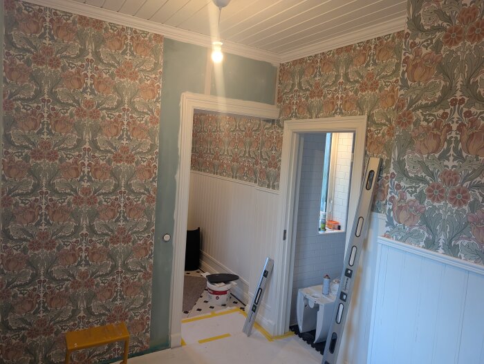 Rum under renovering med färgglada blommiga tapeter på väggarna, en stege och verktyg synliga.