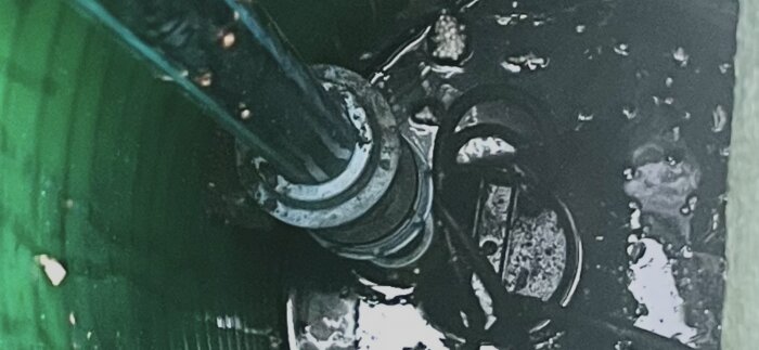 Rör- och kabelinstallation i en mörk, fuktig miljö, delvis under vatten i ett grönt rör eller brunn.