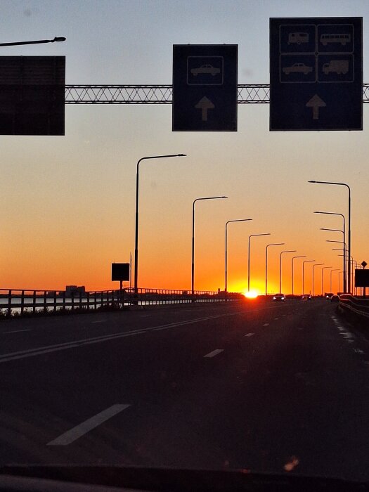 Motorväg mot solnedgången med vägskyltar ovanför och gatlampor längs vägen.