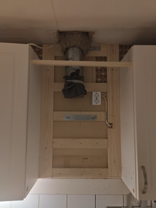 En byggd träram inbyggd mellan köksskåpen med ett delat stänkskydd, exponerade kablar, ett utlopp och en fläktkanal med isolering.