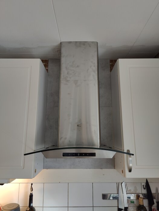Köksfläkt installerad mellan två vita skåp, med påbörjat arbete på Ikea-stänkskydd och ofogade kanter synliga.