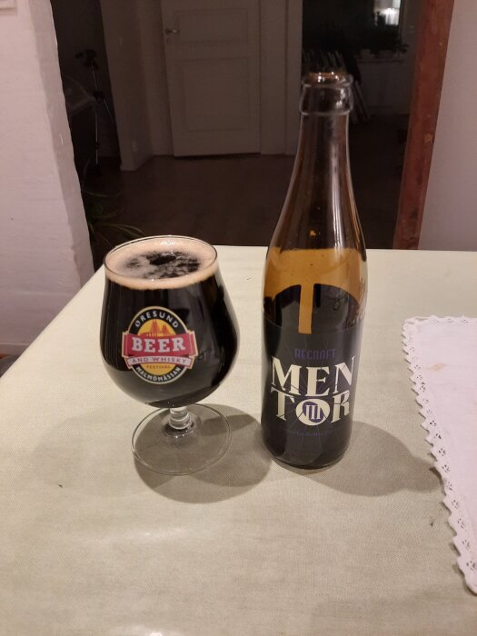 Ölflaska och ölglas på bord, flaskan märkt "Mentor" och glaset med "Beer and Whisky Festival" logga.