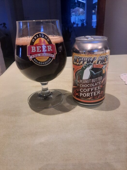 Ett glas med mörk porter på duk, bredvid en burk Hoppin' Frog Peanut Butter Chocolate Coffee Porter.