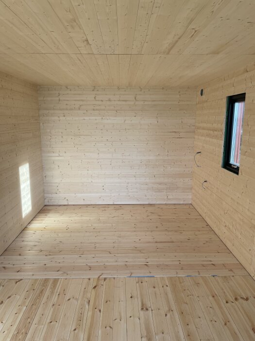 Väggar och golv i ett rum med nyligen monterad träpanel, inga synliga skarvar och ett fönster till höger.
