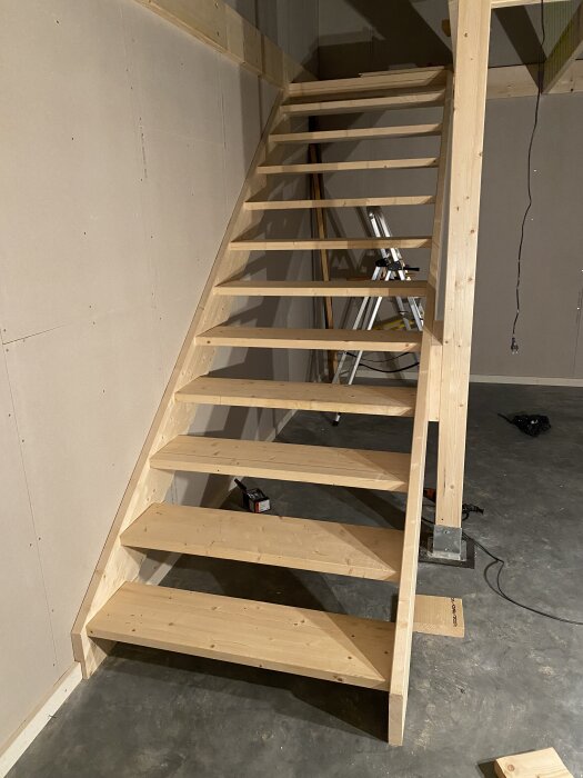 Trätrappa under konstruktion med plywoodsteg i ett rum med betonggolv och en stege i bakgrunden.