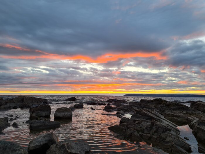 Soluppgång vid havet med klippor i förgrunden och dramatiska moln på himlen.