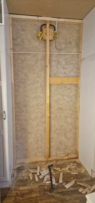 En väggöppning utan väggbeklädnad, med träreglar och synliga elkablar. Spånrester och en kofot ligger på golvet.